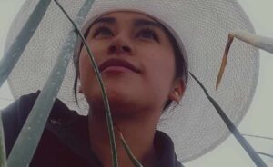 Imagen inspiradora de una joven líder rural entre hojas verdes, con sombrero blanco que simboliza trabajo y protección. Ideal para contenidos sobre empoderamiento femenino, liderazgo agrícola y sostenibilidad en zonas rurales.