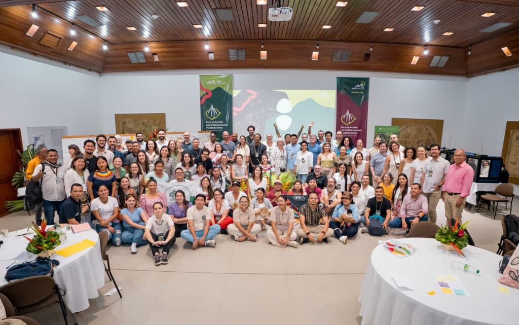 Grupo de participantes reunidos en un evento institucional en un salón amplio con techo de madera y excelente iluminación. La fotografía muestra una escena colaborativa con personas de pie y sentadas, mesas decoradas con manteles blancos y arreglos florales, y dos banners visibles que representan a las organizaciones anfitrionas. Ideal para contenidos sobre encuentros comunitarios, talleres participativos o actividades de fortalecimiento organizacional.